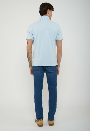 Uomo con tatuaggio al braccio che indossa una polo azzurro chiaro, jeans blu e scarpe da ginnastica beige, in piedi di spalle su uno sfondo semplice.