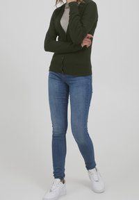 Zelený pletený kardigan s výstřihem do V a zapínáním na knoflíky, doplněný modrými úzkými džíny a bílými teniskami na neutrálním pozadí.