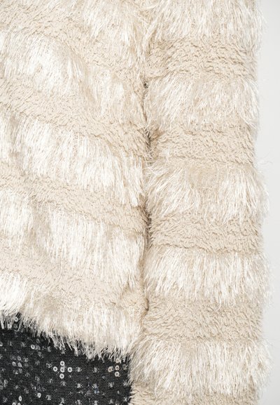 Cremefarbener, fuzzy Pullover mit horizontalen strukturierten Fransenstreifen, getragen mit schwarzen Pailletten-Hosen.
