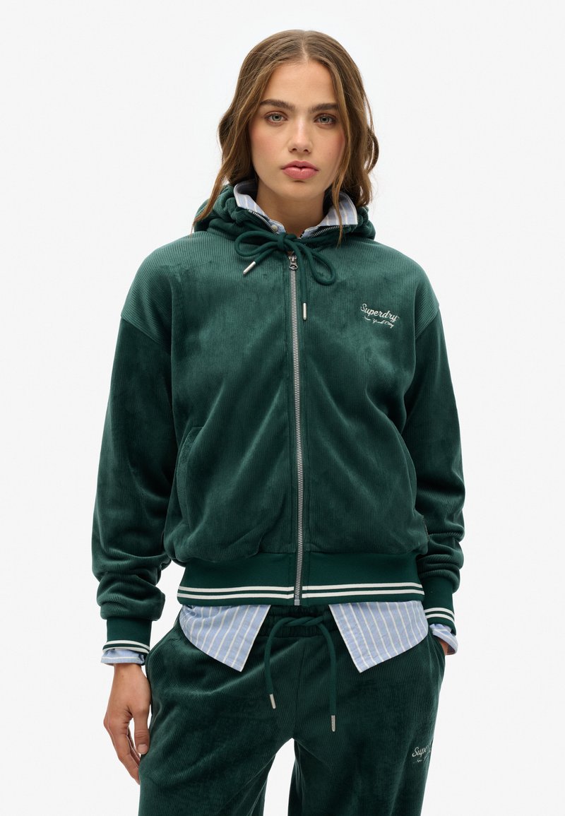 Felpa verde in velluto con zip, dotata di cordino, polsini a costine e vita con strisce bianche. Abbinata a una camicia a righe blu chiaro sotto.