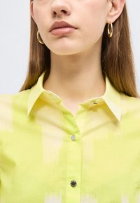 Nő világos sárga, galléros, gombos ingben és arany karika fülbevalókkal, egyenes barna hajjal, amely a vállán túl ér.