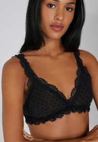 Bralette de encaje negro con lunares, con un diseño triangular y bordes en forma de ondas en las tirantes y el dobladillo. Textura semitranslúcida.