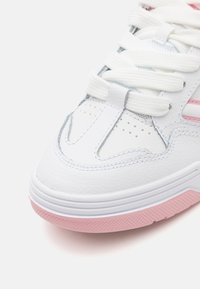 Zapatilla de cuero blanca con suela de goma rosa. Las características incluyen puntera perforada, cordones blancos y detalles rosas en los laterales. Textura suave.