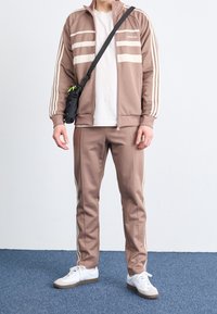 Tuta marrone con strisce bianche sulla giacca e sui pantaloni, realizzata in tessuto liscio; abbinata a una t-shirt bianca e a sneakers bianche.