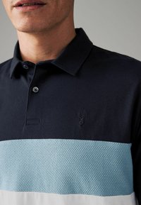 Tumed polo särk, millel on helesinine tekstuuriga riba ja valge alumine riba. Särgil on rinnal tagasihoidlik broderitud hirve logo.