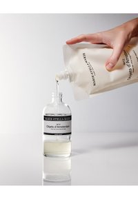 Bouteille de liquide pour les mains en verre transparent avec une étiquette noire, remplie d'un liquide clair, en train d'être remplie à partir d'un sachet crème.
