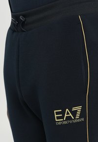 Tmavomodré tepláky s nastaviteľným pásom, s kultovým logom EA7 v zlatej farbe a žltým lemovaním po stranách nohavíc. Hladká textúra.