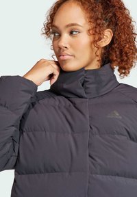 Czarna kurteczka puchowa z wysokim kołnierzem, z pikowanymi sekcjami. Logo Adidas w subtelnym, odbijającym świetle wzorze. Gładka tekstura materiału.