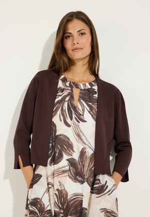 Frau trägt eine braune Jacke mit offenem Vorderteil über einem weißen Kleid mit großen braunen Blumenmustern und einem kleinen Schlüsselloch-Ausschnitt vorne.