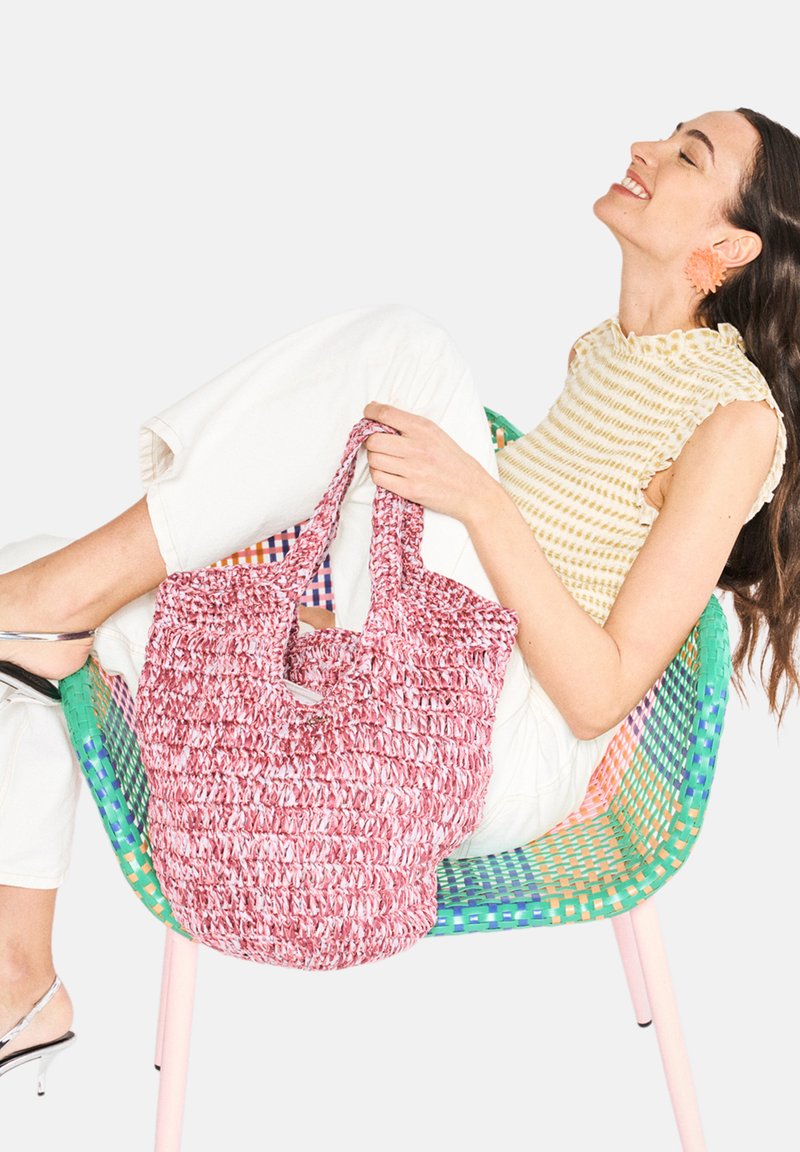 Röd och vit vävd tote-väska med två handtag; strukturerad tyg; hålls av en person som sitter på en färgglad, mönstrad stol.
