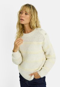 Pull en tricot crème avec des motifs texturés, présentant des rayures horizontales et des embellissements sur les épaules. Tissu texturé et confortable.