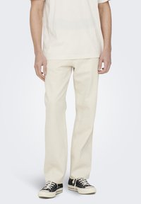 Beige straight-leg pants made of cotton, featuring a smooth texture. Paired with black sneakers and a light beige t-shirt.