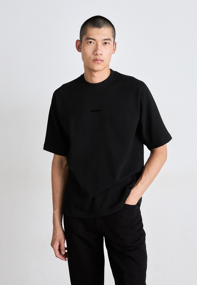 Schwarzes T-Shirt mit kurzen Ärmeln aus weichem Stoff, mit Rundhalsausschnitt und einem dezenten horizontalen Nahtdetail über der Brust. Kombiniert mit dunklen Hosen.