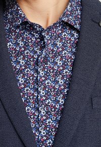 Närbild av en person som bär en marinblå blommig skjorta med vita, röda och blå blommor under en strukturerad mörkblå kavaj.