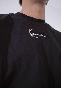 Svart t-skjorte laget av bomull, med en hvit brodert signaturlogo på brystet. Crew neck-design, glatt tekstur.