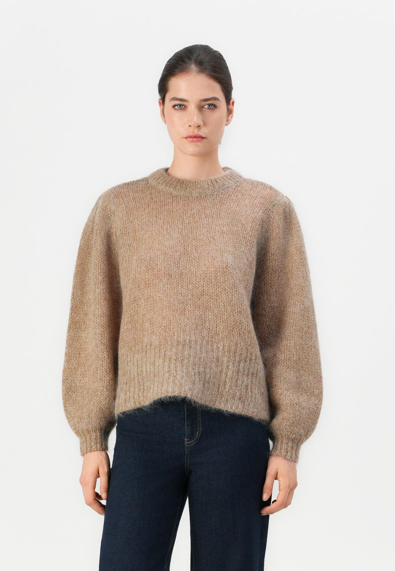 Maglione di colore marrone chiaro in maglia con scollo rotondo e maniche a palloncino. Tessuto morbido e texturizzato con orlo a costine, abbinato a jeans scuri.