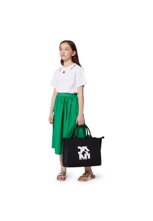 Zwarte tote bag met wit logo, gedragen door een kind in een witte shirt met uitsparingen, groene rok en gouden schuifslippers. Minimalistisch ontwerp.