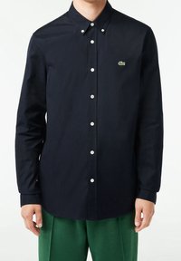 Camicia blu navy con bottoni realizzata in tessuto dalla texture liscia. Presenta un colletto a punta, bottoni bianchi e un piccolo logo verde sul petto.