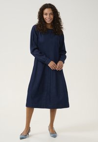 Donkerblauwe denim jurk met lange mouwen, een ontspannen pasvorm, een ronde halslijn en gathered mouwen, tot halverwege de kuit reikend. Gepaard met lichtblauwe hakken.