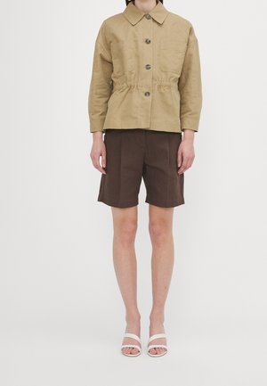 Osoba ubrana w beżową kurtkę zapinaną na guziki z przednimi kieszeniami, ciemnobrązowe krótkie spodenki do kolan oraz białe sandały z odkrytymi palcami, stojąca na jednolitym tle.