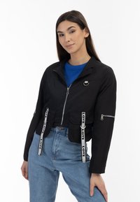 Giacca nera corta con zip frontale, dettagli argentati, cinturini con stampa Love e colletto rialzato. Abbinata a un top blu e jeans.