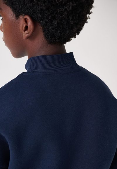 Tumši zils džemperis ar augstu ribotiem apkakli. Gluda, teksturēta auduma ar pieguļošu dizainu. Minimālistisks stils, bez redzamām rakstām vai logotipiem.