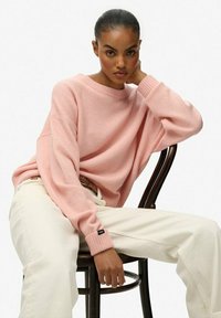 Rosa Strickpullover mit legerem Schnitt und gerippten Bündchen, kombiniert mit cremefarbenen Hosen. Das Modell sitzt auf einem Holzstuhl vor einem schlichten Hintergrund.