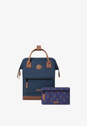 Tummansininen kangaskassi, jossa on ruskeat nahkakiinnitykset ja etupuolella vetoketjutasku. Paketti sisältää yhteensopivan kuvioidun vetoketjupussin.