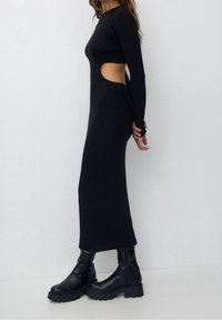 Robe noire à manches longues avec découpe latérale, coupe ajustée et tissu doux. Associée à des bottines noires à semelle épaisse. Design minimaliste.