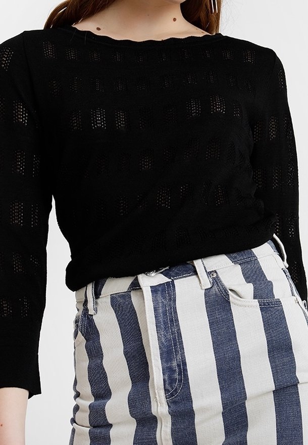 Femme portant un haut noir à manches longues en maille avec un motif à carreaux et une jupe taille haute blanche à rayures verticales bleues.