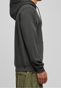 Tummanharmaa huppari, jossa on kiristysnyörillä varustettu huppu, kengurutasku ja resorit hihansuissa. Pehmeä pinta ja rento istuvuus, yhdistetty vihreisiin cargo-housuihin.