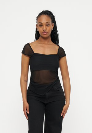 Γυναίκα με μακριά πλεξούδα φοράει μαύρο διαφανές μπλουζάκι με κοντά μανίκια και μαύρο παντελόνι, στέκεται με ουδέτερη έκφραση μπροστά σε λευκό φόντο.