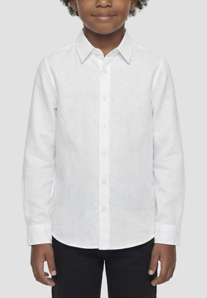 Dijete odjeveno u bijelu košulju s dugim rukavima i crne hlače, stoji ispred jednobojne svijetlosive pozadine.