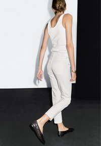 White tank top and light gray trousers, with a fitted design. Black flat shoes feature a textured finish. Background is monochrome.
