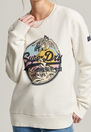 Osoba nosząca biały sweter z kolorowym logo Superdry przedstawiającym żurawia i wzór fali, zestawiony z niebieskimi dżinsami.