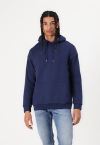 Navy blue hooded sweatshirt made of soft cotton blend, featuring a front pocket and adjustable drawstring. Model wearing light blue jeans.