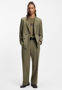 Olivgrüner, maßgeschneiderter Anzug mit einem doppelseitigen Blazer, strukturiertem Stoff, locker geschnittenen Hosen und einem passenden olivgrünen T-Shirt mit schwarzem Logo.