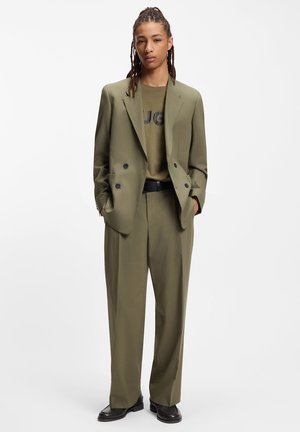 Olivgrüner, maßgeschneiderter Anzug mit einem doppelseitigen Blazer, strukturiertem Stoff, locker geschnittenen Hosen und einem passenden olivgrünen T-Shirt mit schwarzem Logo.