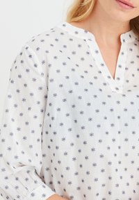 Blusa bianca con un sottile motivo geometrico navy, scollatura a V e maniche a tre quarti con dettagli ai polsini. Tessuto leggero e testurizzato.