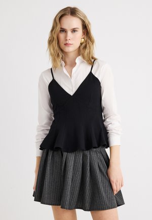 Ung kvinde med bølget blondt hår iført guldøreringe, hvid skjorte med knapper, sort ærmeløs peplumtop og grå nederdel med striber.