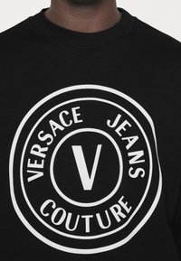 Schwarzer Pullover mit einem großen, weißen kreisförmigen Logo, das den Text "VERSACE JEANS COUTURE" und ein zentrales, fett gedrucktes "V" zeigt. Weiche Stoffstruktur.