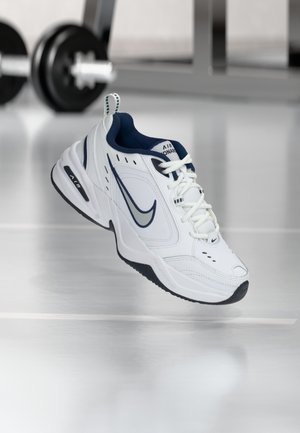 Baskets Nike Air Monarch blanches et bleu marine flottant au-dessus d'un sol réfléchissant avec des poids de gym flous en arrière-plan.