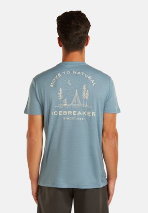 Hombre con camiseta azul que lleva un gráfico en la espalda con una fogata, árboles, luna y el texto "MOVE TO NATURAL ICEBREAKER SINCE 1995".