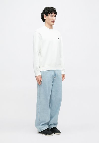 Λευκό φούτερ με στρογγυλή λαιμόκοψη και μικρό λογότυπο, συνδυασμένο με ανοικτόχρωμο φαρδύ τζιν και μαύρα αθλητικά παπούτσια. Ομοιόμορφη υφή υφάσματος.