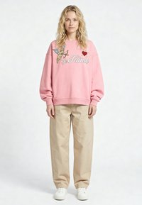 Femme blonde portant un sweat-shirt rose avec un cupidon et le texte "Je T'aime", un pantalon beige à jambes larges et des baskets blanches, debout devant un fond uni.