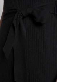 Tessuto nero a coste con un dettaglio di fiocco legato in vita. La texture è morbida ed elastica, evidenziando una silhouette aderente.