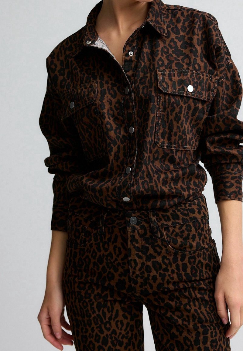 Femme porte une chemise boutonnée imprimé léopard marron et noir avec un pantalon assorti, montrant les détails de la texture du tissu et des boutons.