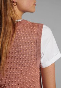 Stickad ärmlös topp i mjuk rosa med strukturerat prickmönster. Har en ribbad halsringning och fåll, lagd över en vit t-shirt.