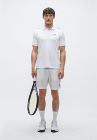 Hvit atletisk poloshirt med korte ermer, lysegrå shorts og hvite atletiske sko; holder en tennisracket. Enkel, funksjonell design.