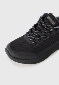 Juodi Skechers BOBS sportbačiai su tekstūruota viršutine dalimi, raštuotais juodais ir pilkais raiščiais bei paminkštintu padu ant balto fono.